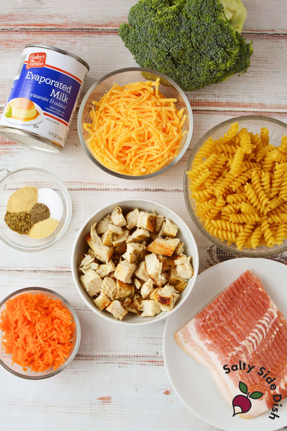 Ingredients for broccoli cheese chicken pasta, including chicken, bacon, broccoli, carrots, rotini, cheddar cheese, evaporated milk, and seasonings.