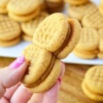 A hand holding two homemade copycat Nutter Butter sandwich cookies with a plate of cookies in the background