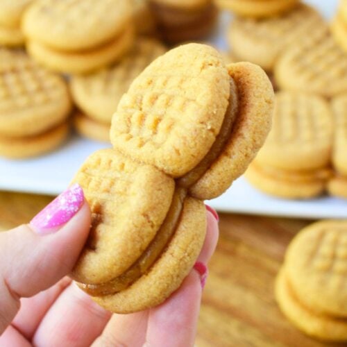 A hand holding two homemade copycat Nutter Butter sandwich cookies with a plate of cookies in the background