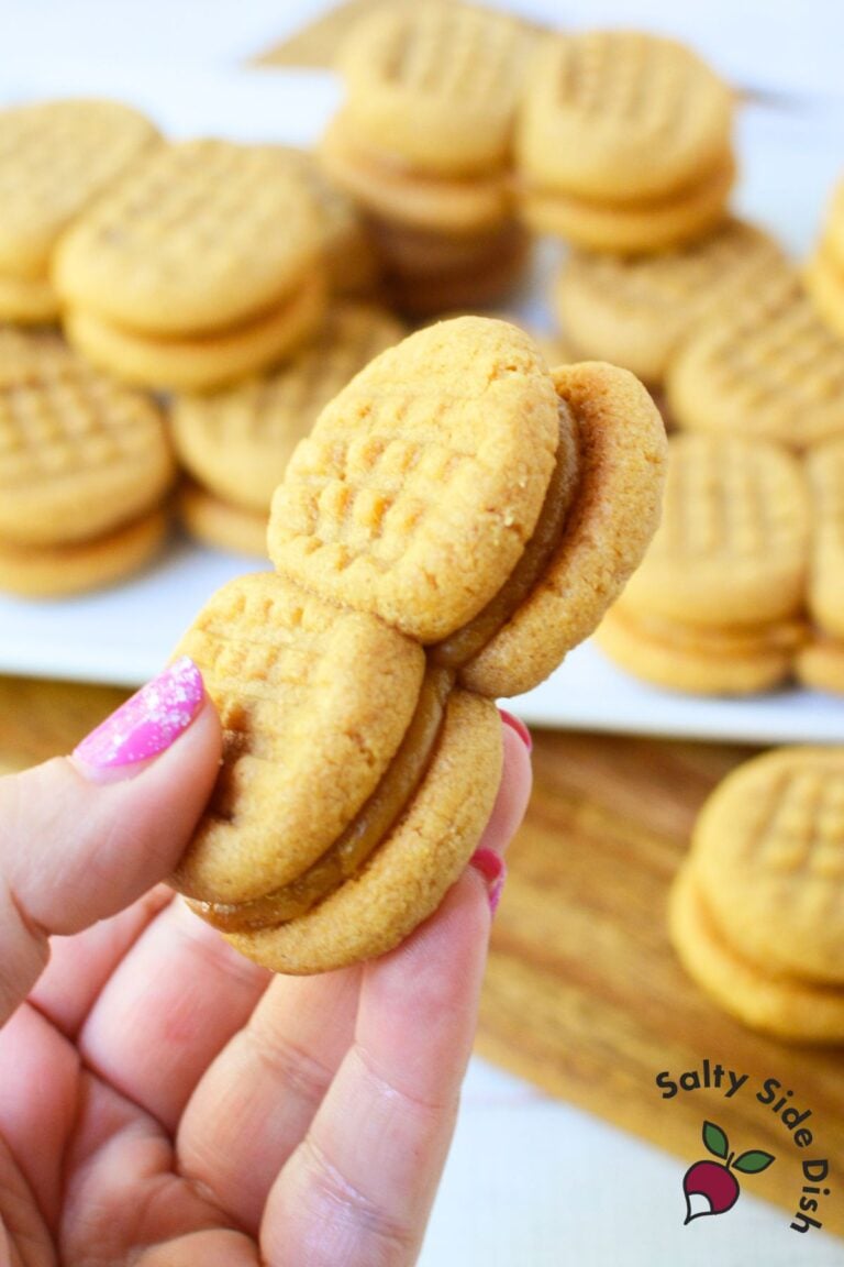 A hand holding two homemade copycat Nutter Butter sandwich cookies with a plate of cookies in the background