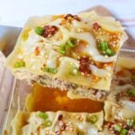 A slice of pork dumpling lasagna topped with chili oil, sesame seeds, and sliced green onions.