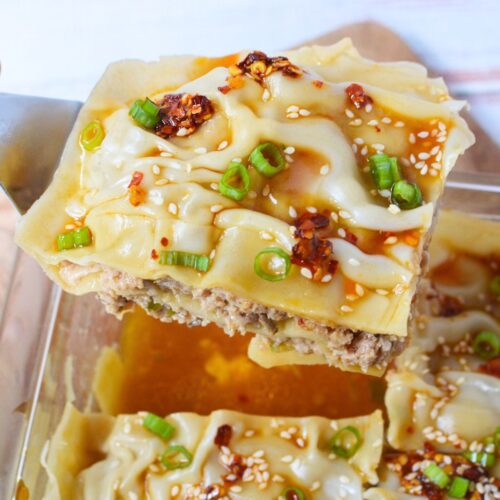 A slice of pork dumpling lasagna topped with chili oil, sesame seeds, and sliced green onions.