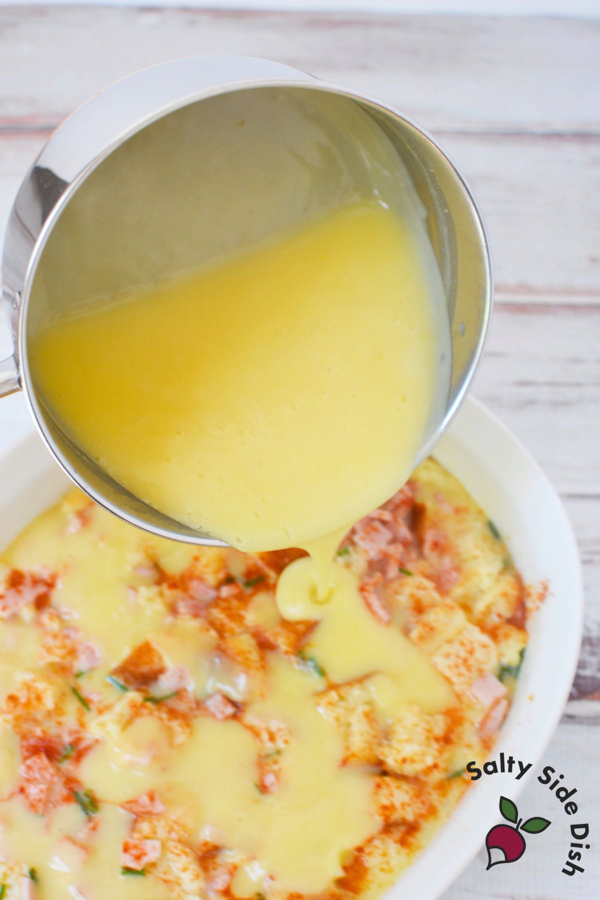 Prepared hollandaise sauce being poured over baked eggs benedict casserole