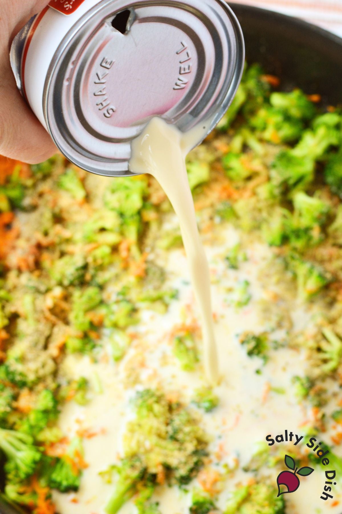 Evaporated milk being poured into a skillet of broccoli, carrots, and seasonings for creamy pasta sauce.
