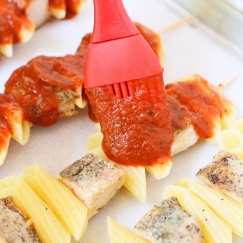 Brushing marinara sauce over chicken and pasta skewers before adding cheese