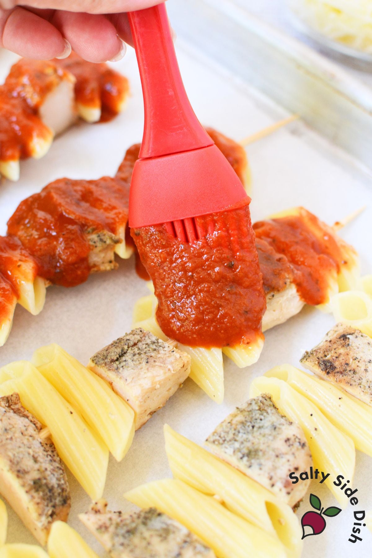 Brushing marinara sauce over chicken and pasta skewers before adding cheese
