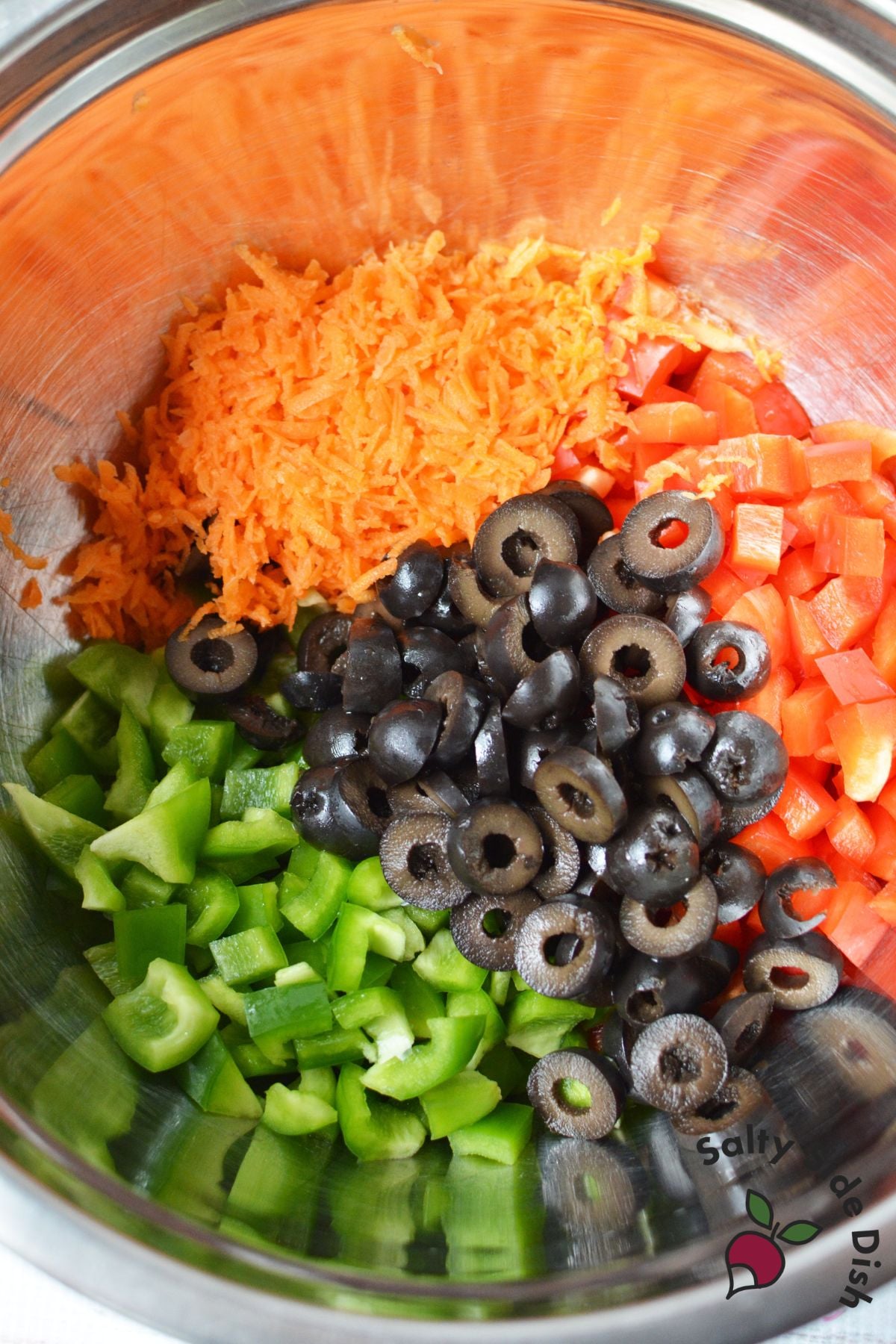 Chopped bell peppers, shredded carrots, and sliced black olives in a mixing bowl for pasta salad