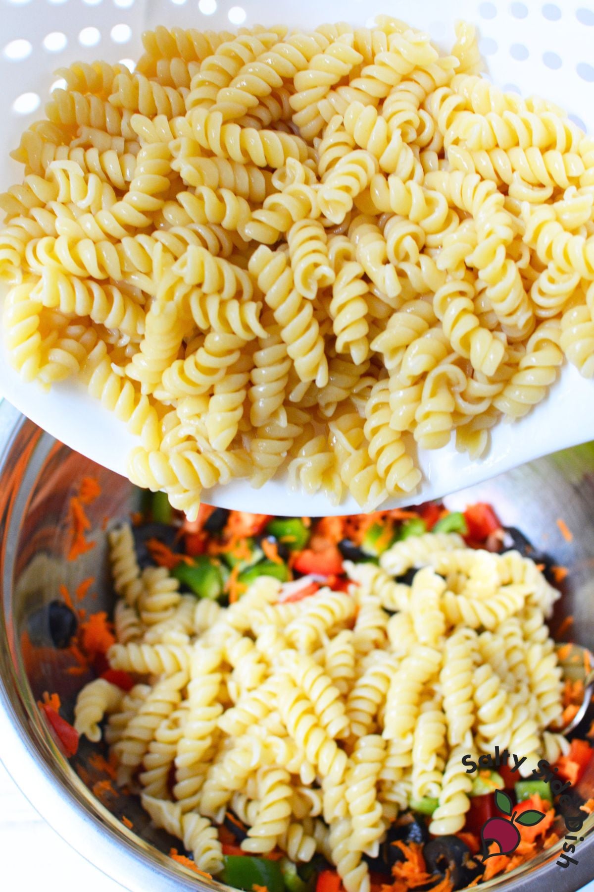 Cooked rotini pasta being added to chopped vegetables and black olives in a mixing bowl