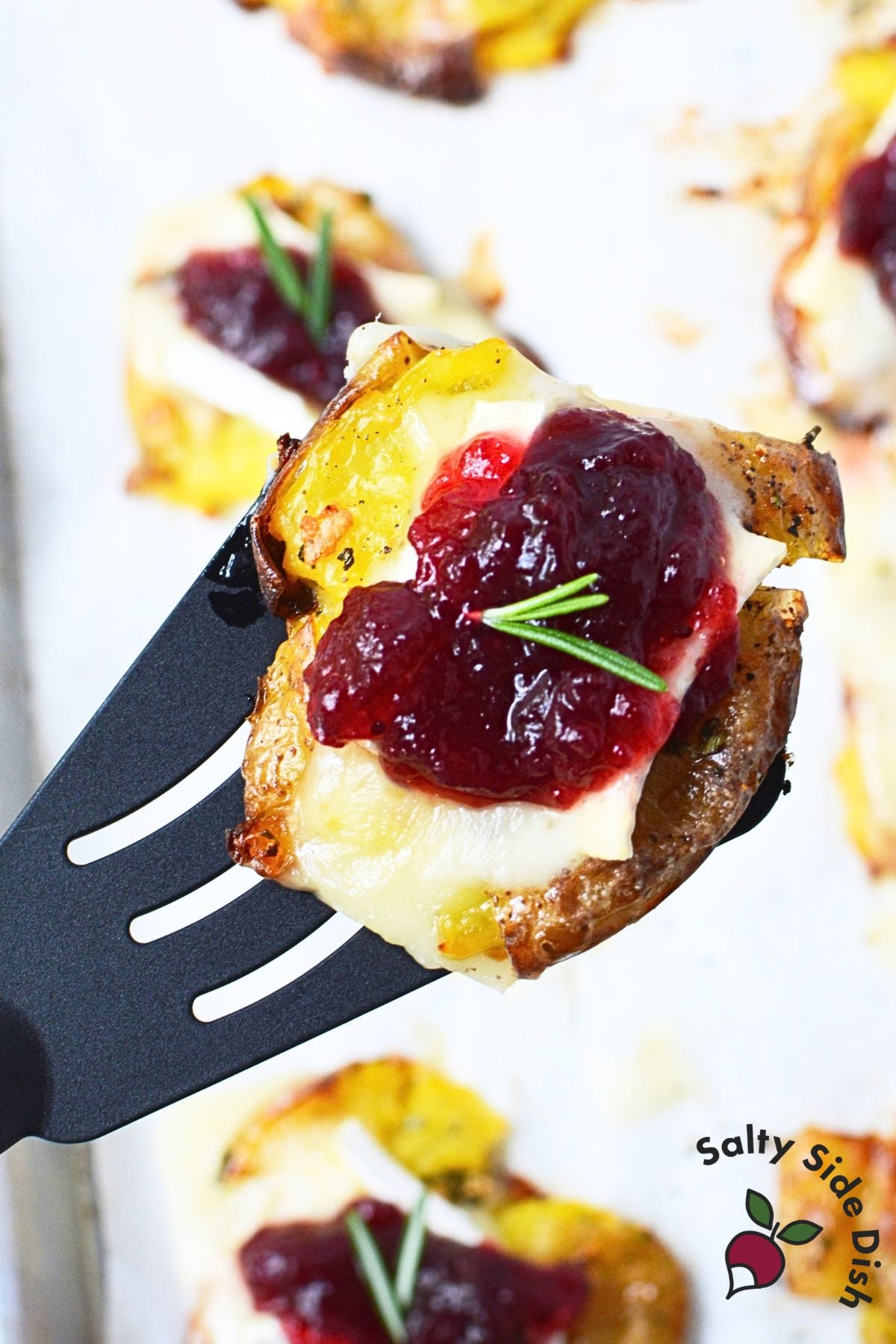 Cranberry Brie smashed potato lifted on a spatula, showing the creamy potato, melted Brie, cranberry sauce, and rosemary topping.