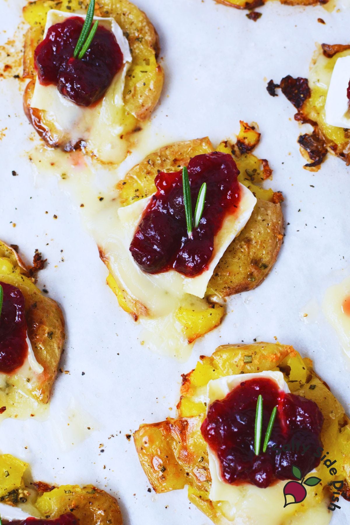 Roasted smashed baby potatoes topped with Brie, cranberry sauce, and fresh rosemary on a baking sheet.