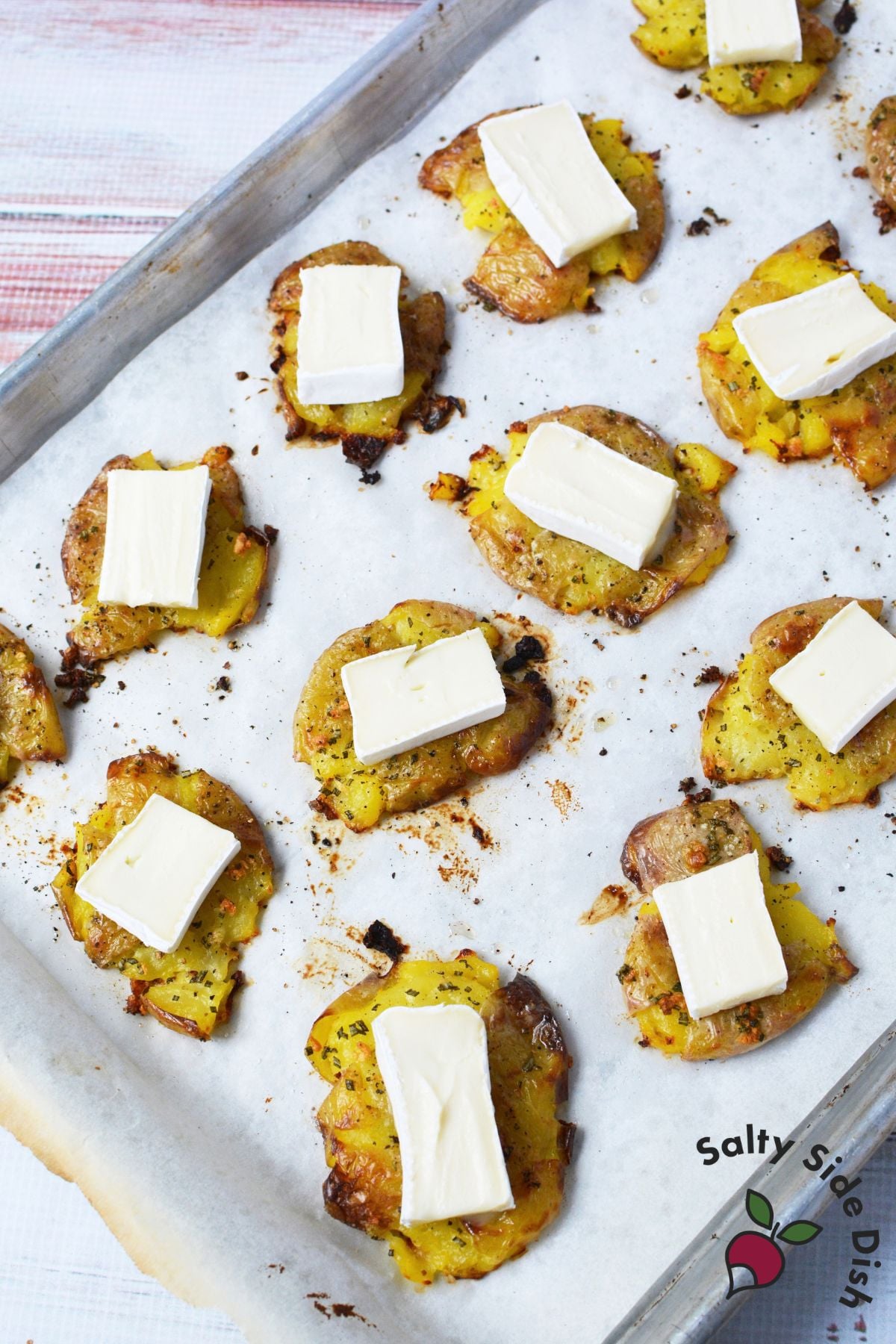 Brie slices added to golden smashed potatoes on a parchment-lined baking sheet before melting.