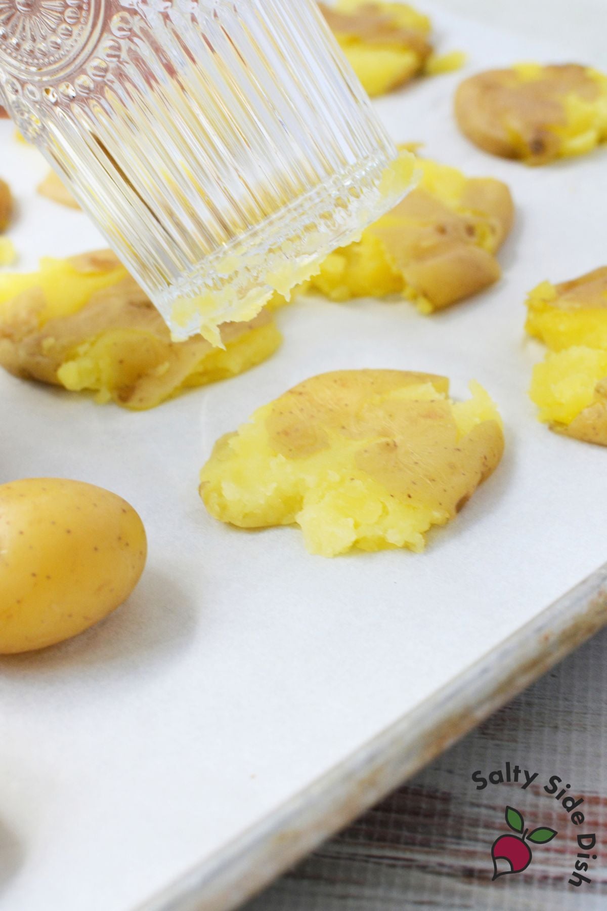 Boiled baby gold potatoes being gently smashed with the bottom of a drinking glass on a baking sheet.