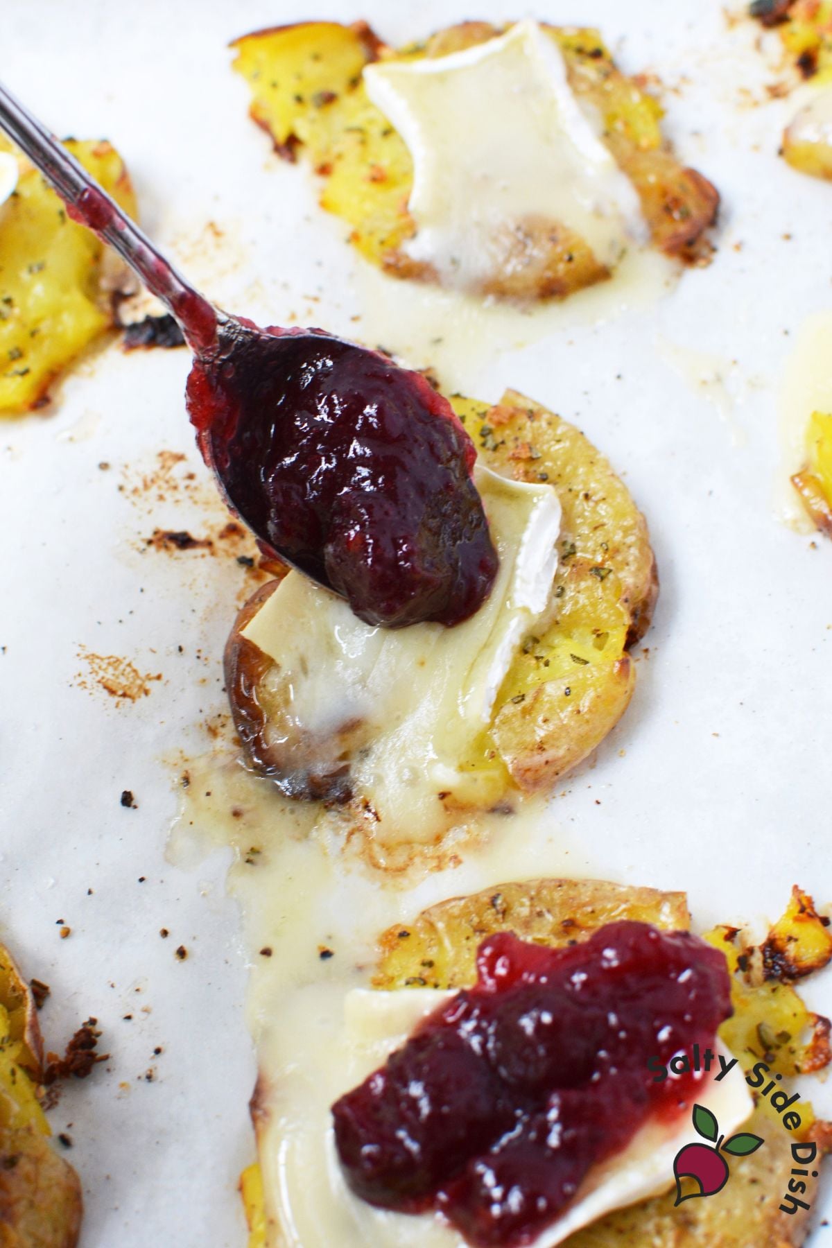 Whole berry cranberry sauce being spooned over melted Brie on roasted smashed potatoes.