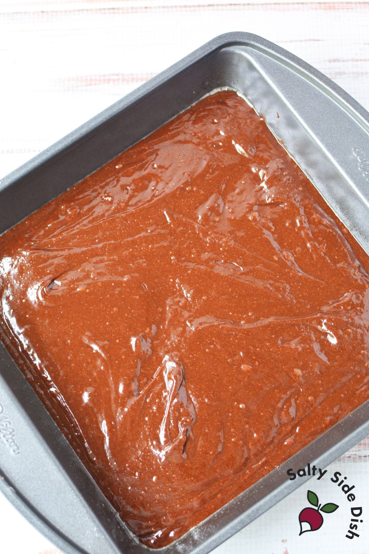Chocolate brownie batter spread into a square baking pan before baking.