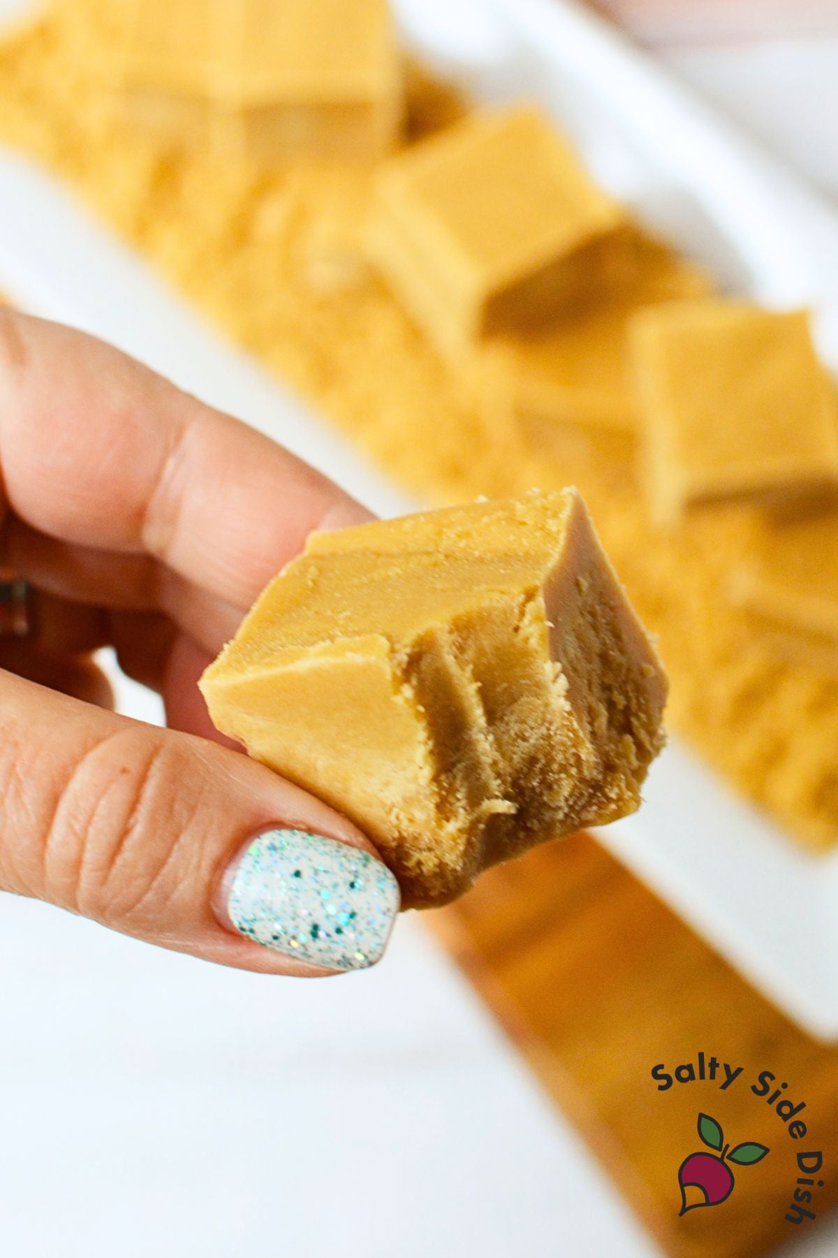 Hand holding a bite-size square of smooth brown sugar fudge with sliced fudge in the background.