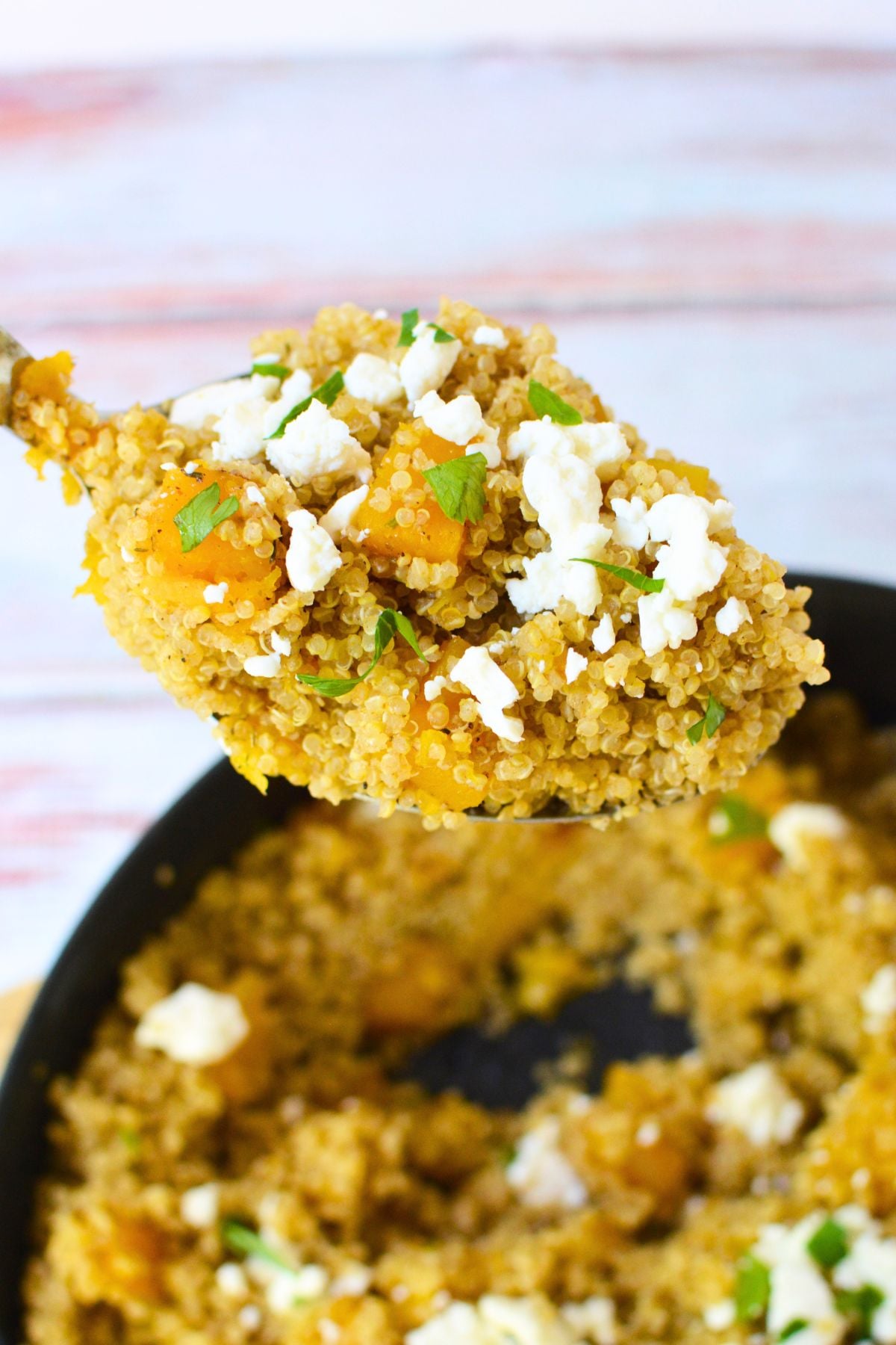 Spoonful of warm butternut squash quinoa salad topped with crumbled feta cheese and chopped parsley above a skillet.