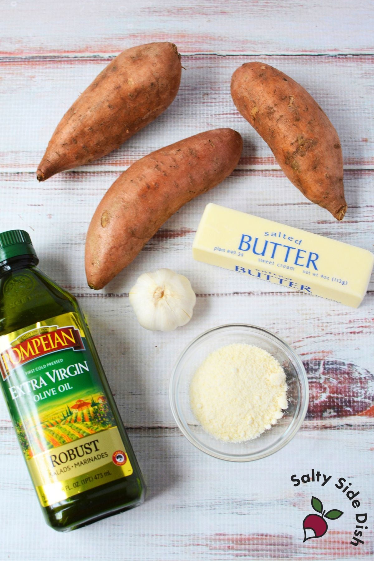 Three whole sweet potatoes with olive oil, butter, garlic, and grated Parmesan arranged on a light wood surface.