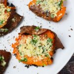 Close-up of a garlic Parmesan smashed sweet potato on a white plate with browned skin, parsley, and black pepper.