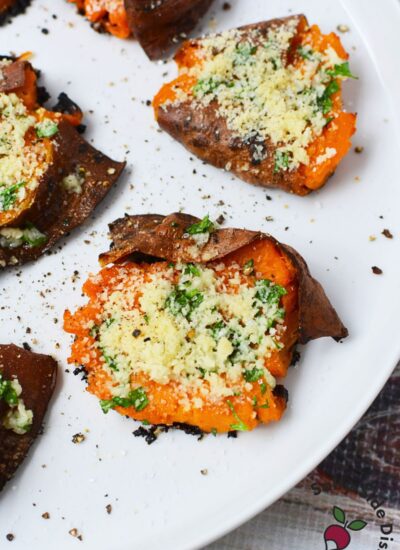 Close-up of a garlic Parmesan smashed sweet potato on a white plate with browned skin, parsley, and black pepper.