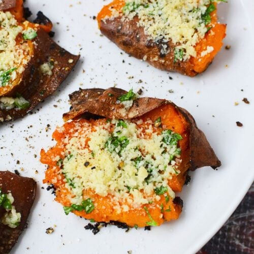 Close-up of a garlic Parmesan smashed sweet potato on a white plate with browned skin, parsley, and black pepper.