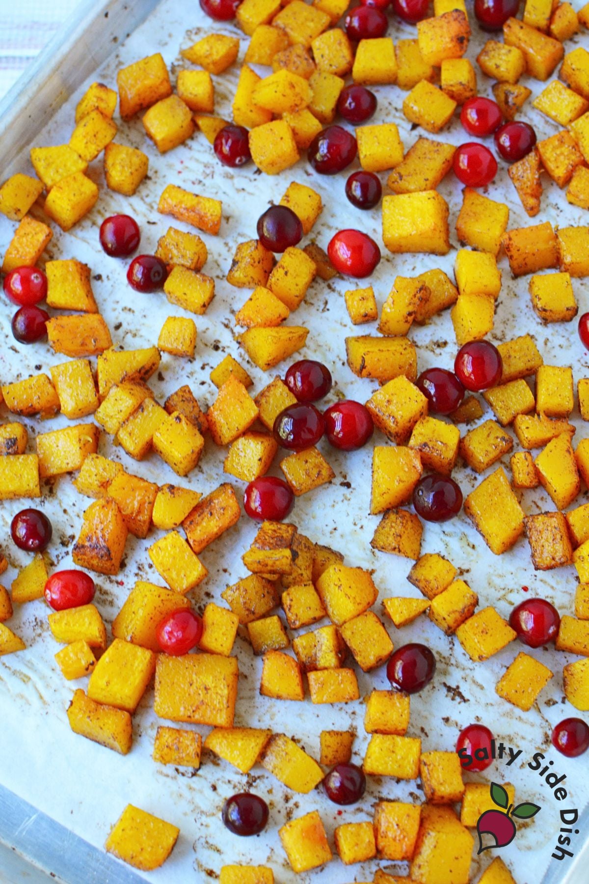 Whole cranberries scattered among butternut squash cubes on a baking sheet.