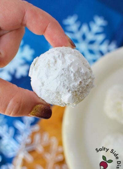 Homemade almond snowball cookie coated in powdered sugar and held between fingers against a blue Christmas snowflake background.