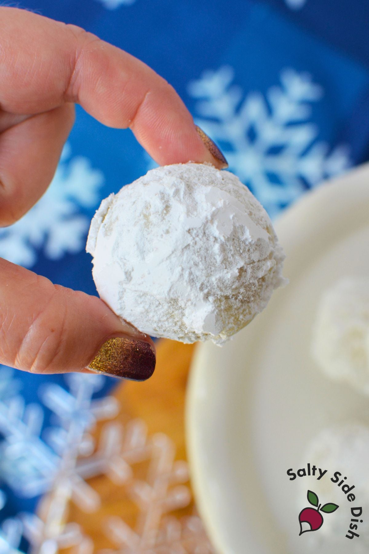 Homemade almond snowball cookie coated in powdered sugar and held between fingers against a blue Christmas snowflake background.