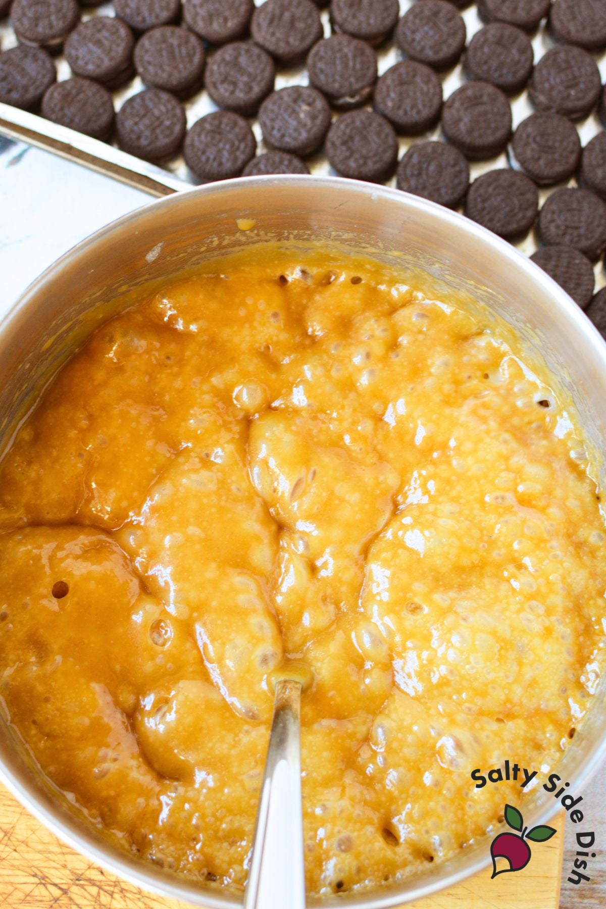 Butter and brown sugar toffee mixture bubbling in a saucepan with Mini Oreo cookies arranged on a baking sheet in the background.