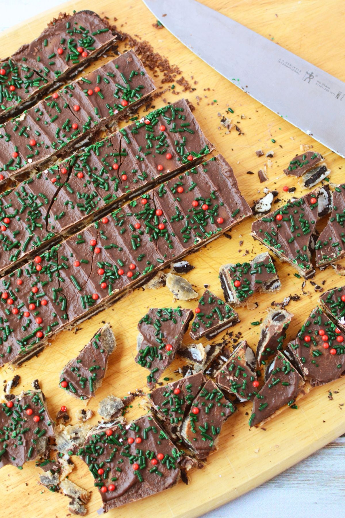 Mini Oreo toffee bark being cut into uneven pieces with a sharp kitchen knife on a wooden cutting board.