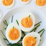Pumpkin deviled eggs with swirled orange filling and green chive stems on a white serving platter