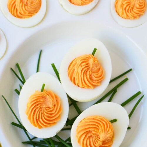 Pumpkin deviled eggs with swirled orange filling and green chive stems on a white serving platter