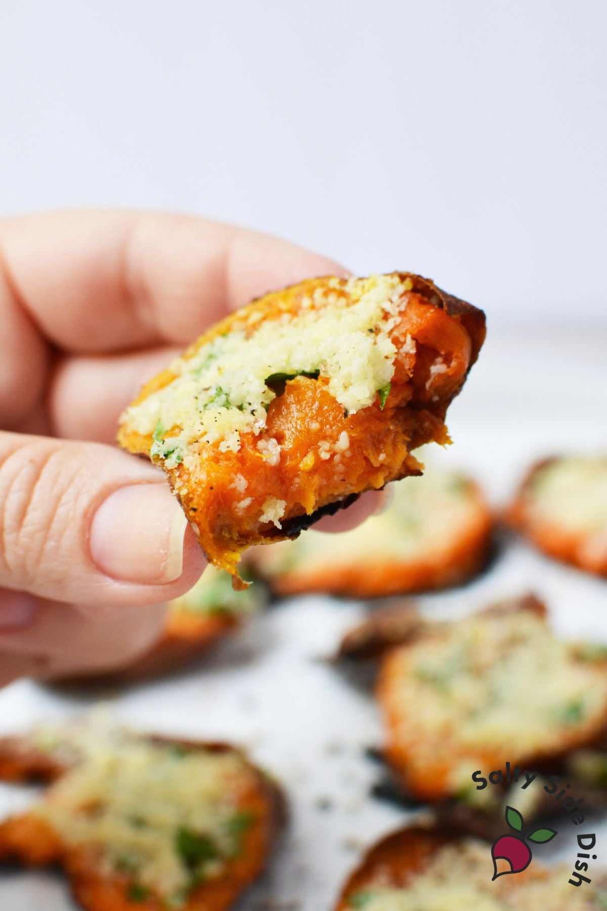 Hand holding a bite of garlic Parmesan smashed sweet potato showing the soft orange center and Parmesan topping.