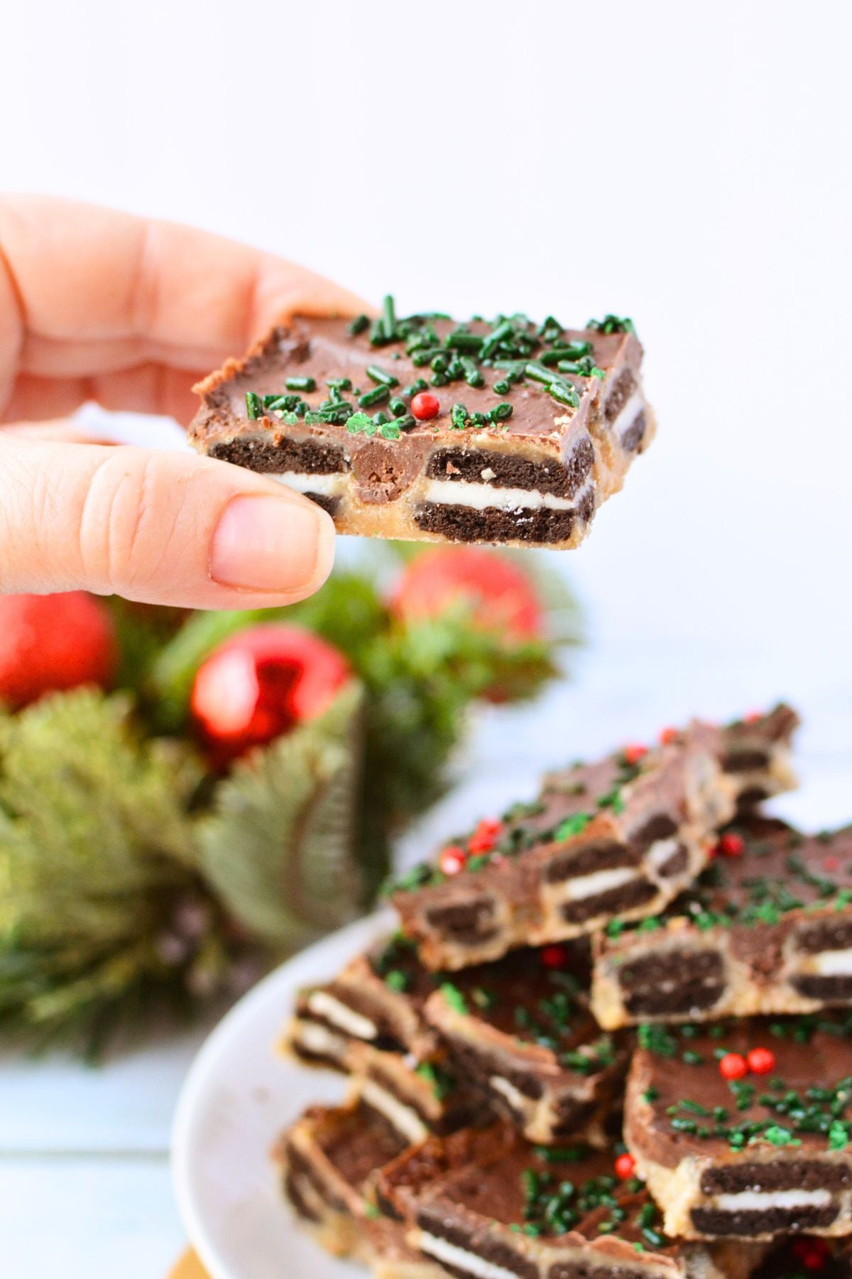 A piece of homemade Mini Oreo Christmas toffee held above a plate of chocolate-covered toffee bark with holiday sprinkles.