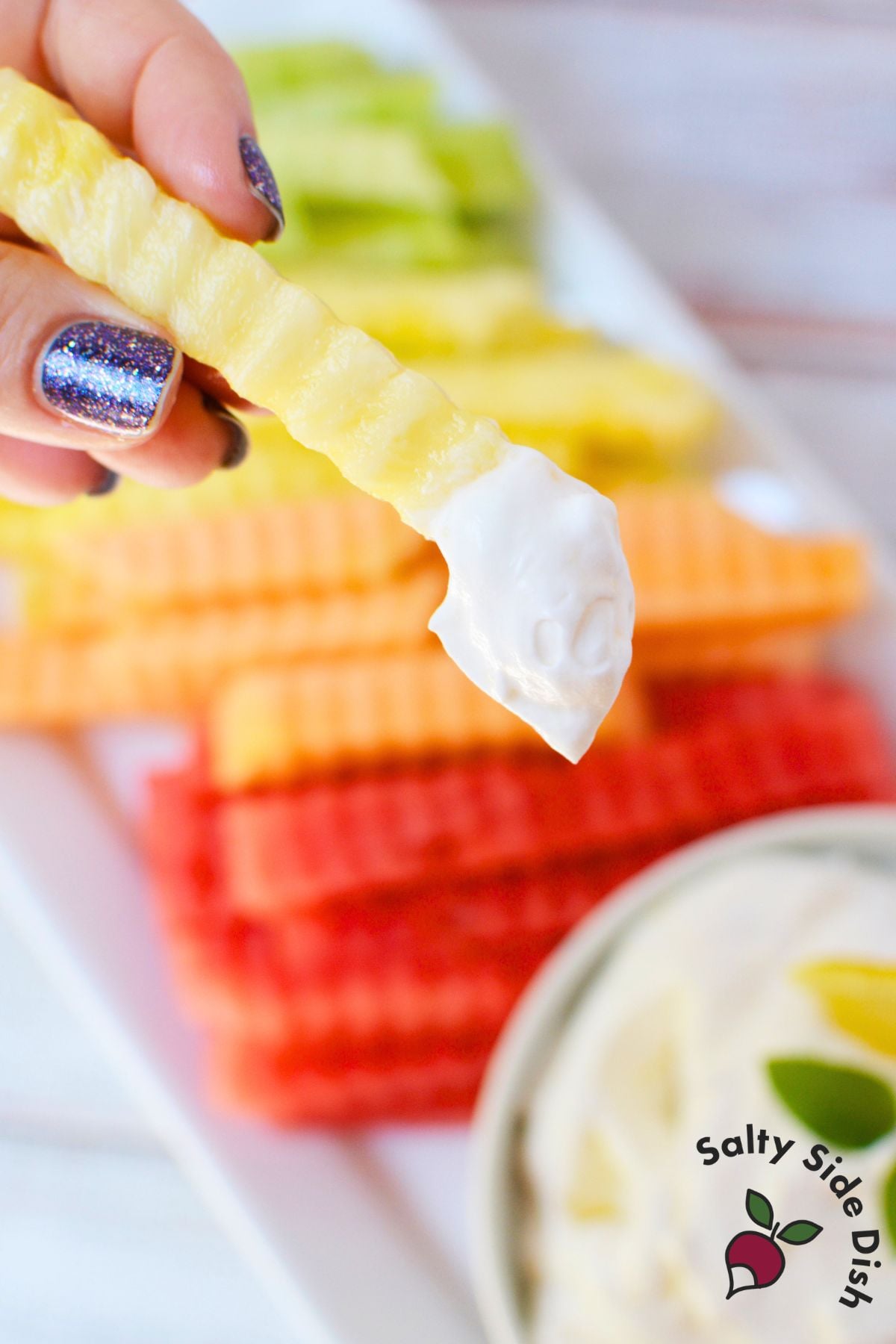 Crinkle-cut pineapple fruit fry dipped into fluffy lemon yogurt dip with colorful melon fries in the background.