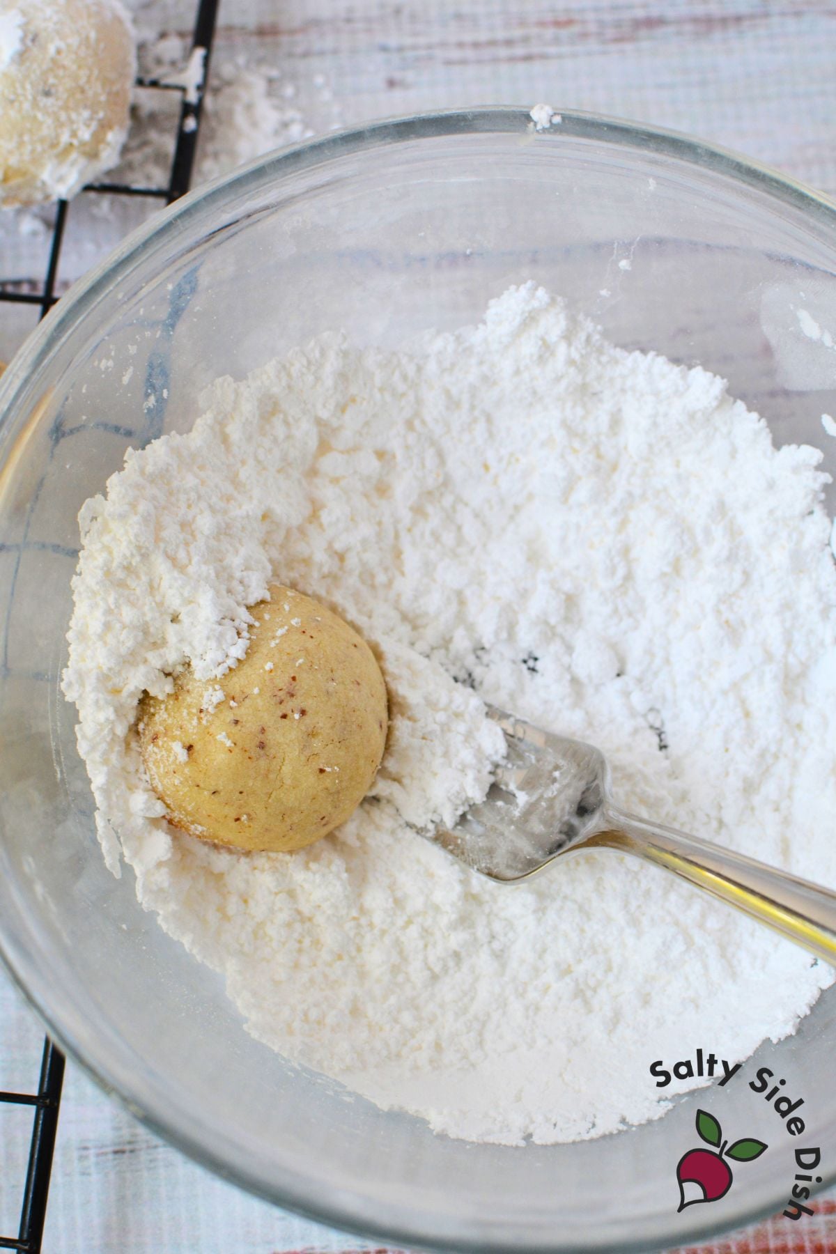Baked almond snowball cookie being rolled in a bowl of powdered sugar for its classic white coating.