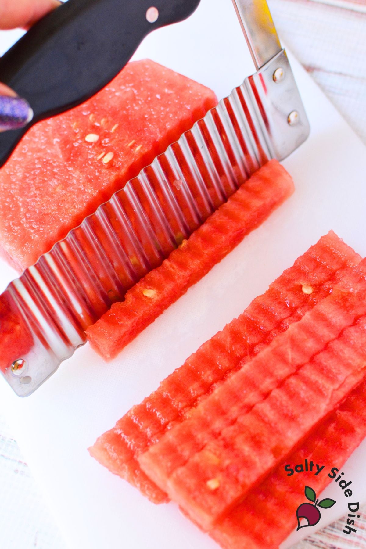 Fresh watermelon fries being cut into wavy strips with a handheld crinkle cutter on a white cutting board.
