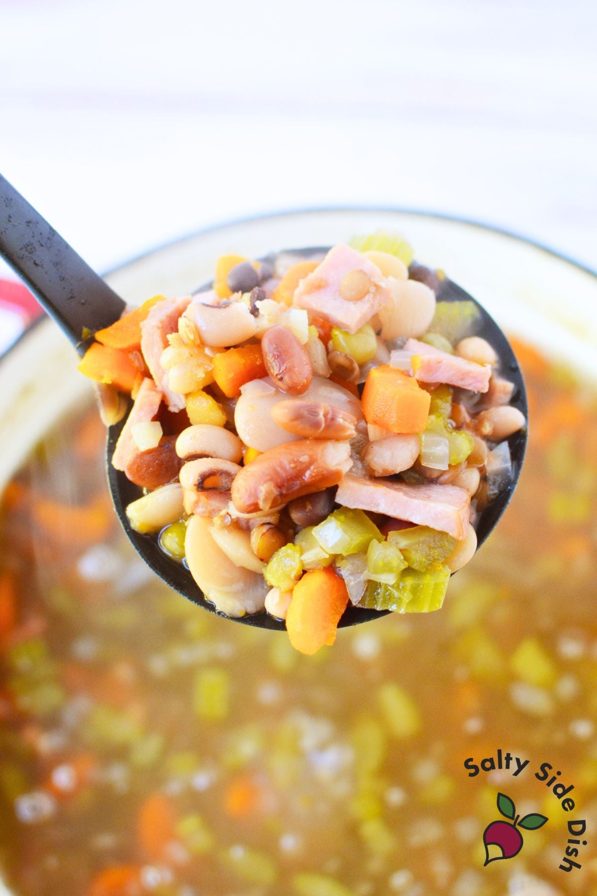 Ladle filled with cooked mixed beans, ham, carrots, and celery above a pot of homemade ham and bean soup.